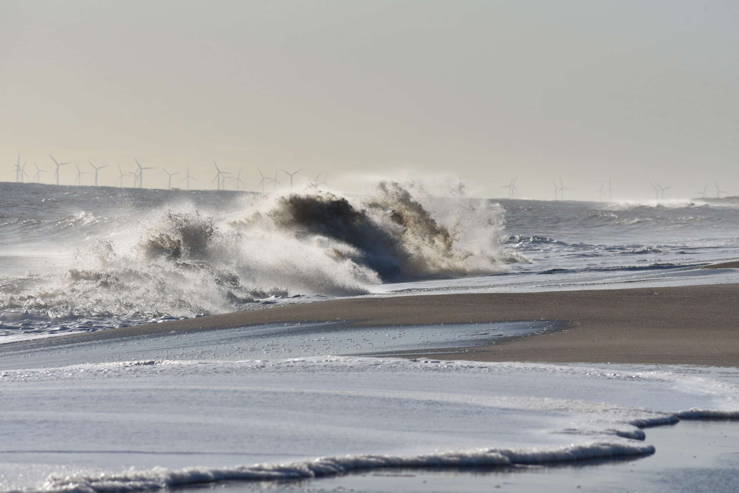 Waves crashing onto a sandy beach with offshore wind turbines on the horizon